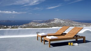 Two wooden lounge chairs with towels and a glass of orange juice on a rooftop terrace overlooking a coastal landscape with white buildings and a mountain in the distance. - Villa Fabrica Santorini (Santorini)