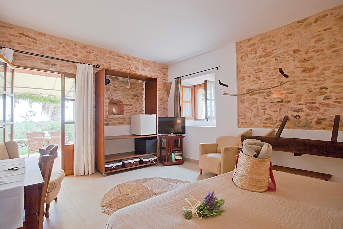 Un dormitorio con pared de piedra, una cama con un ramo, televisor, estante de madera y un rincón comedor con sillas.