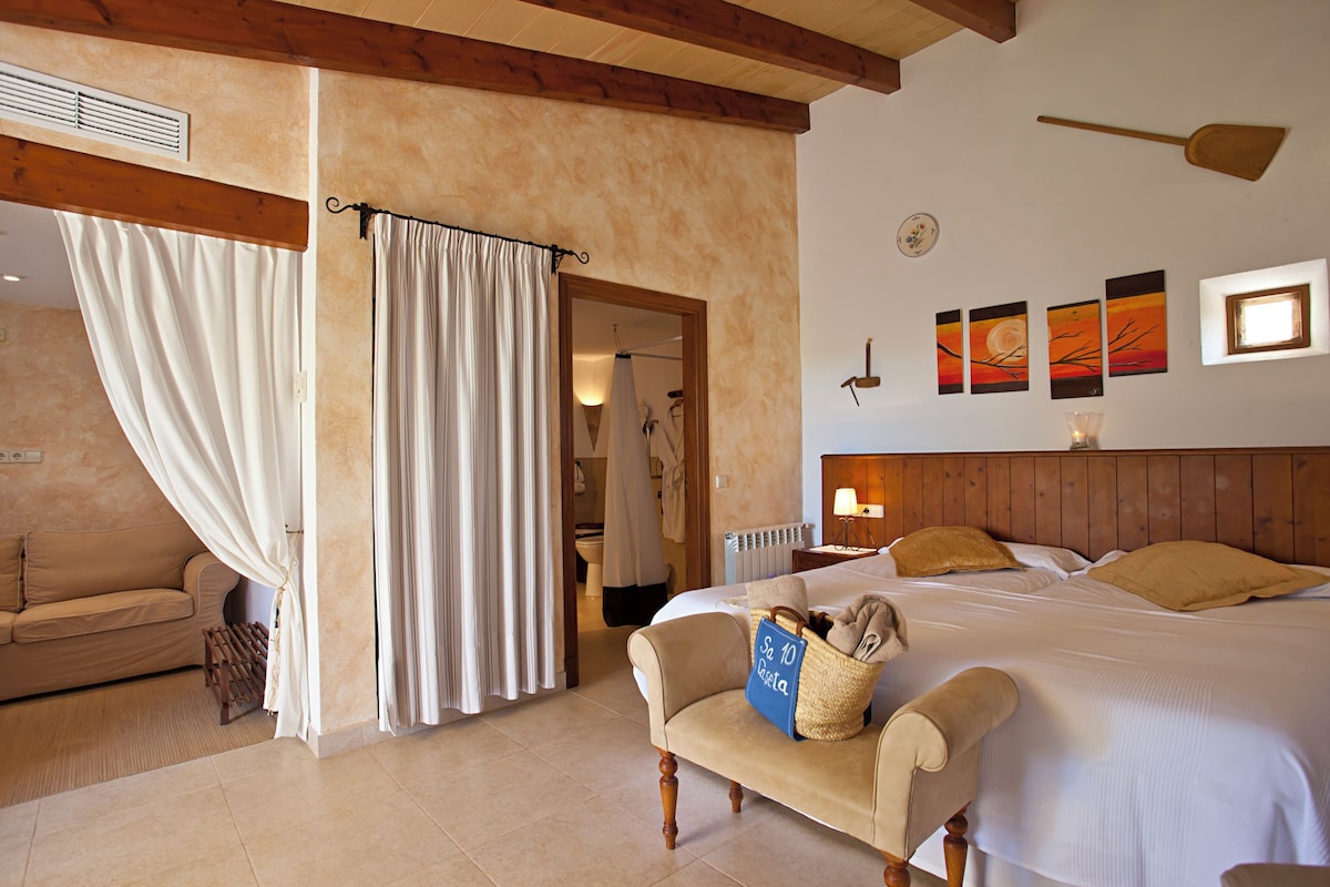 Una habitación de hotel con cama, una silla, una mesita, ventana con cortinas y cuadros en la pared.
