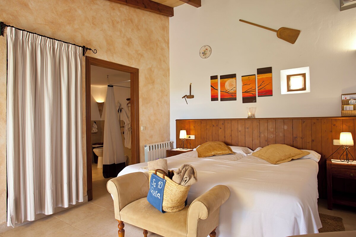 Habitación de hotel con cama, una silla, cabecera de madera, mesita de noche, lámpara y una obra de arte en la pared.