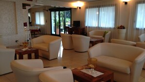 Un vaste salon offrant de nombreuses possibilités d’assise, notamment des canapés et des fauteuils, disposés autour d’une table basse centrale. On y trouve également un téléviseur et une petite table sur laquelle sont posés une lampe et des objets décoratifs. De grandes fenêtres, équipées de rideaux, permettent à la lumière naturelle d’entrer dans la pièce.