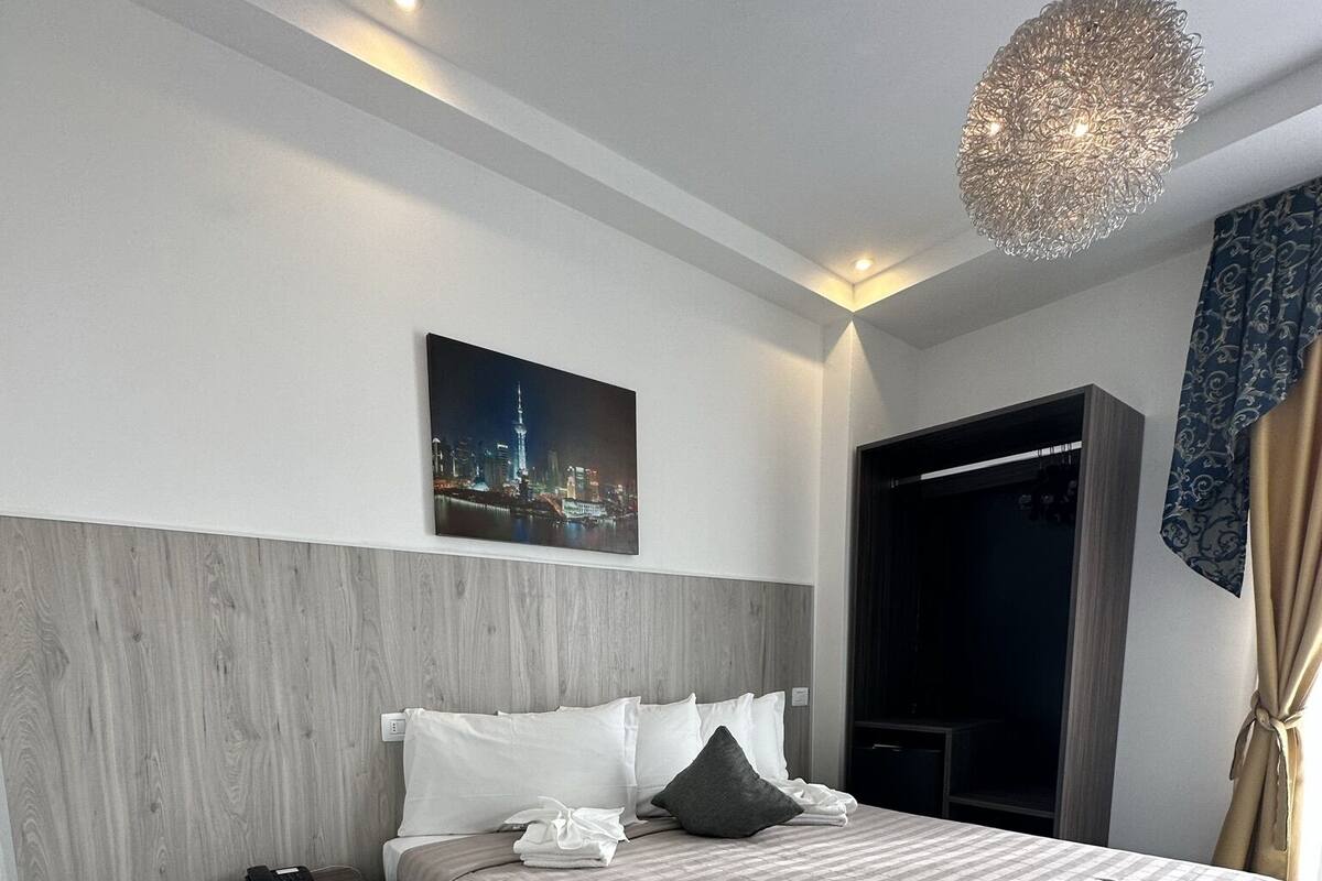 A modern hotel room with a large bed, a nightstand, a telephone, a cityscape painting on the wall, and a unique ceiling light fixture.