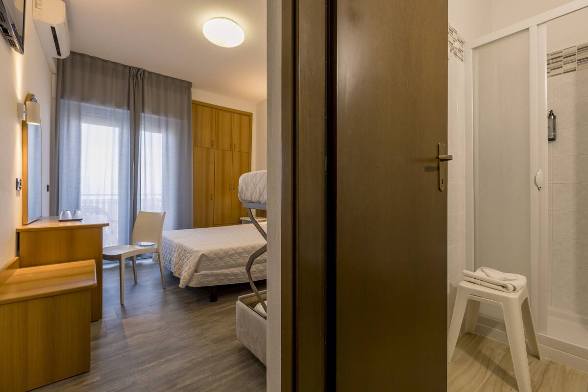 A hotel room with a bed, a desk, a chair, a wardrobe, and a shower area.