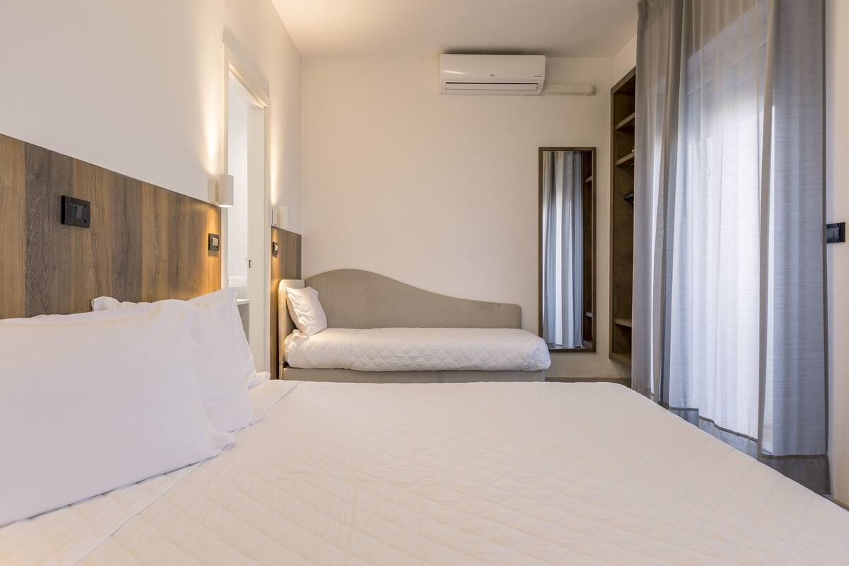 A hotel room with a large bed, a bedside table, a wardrobe, and an air conditioning unit.