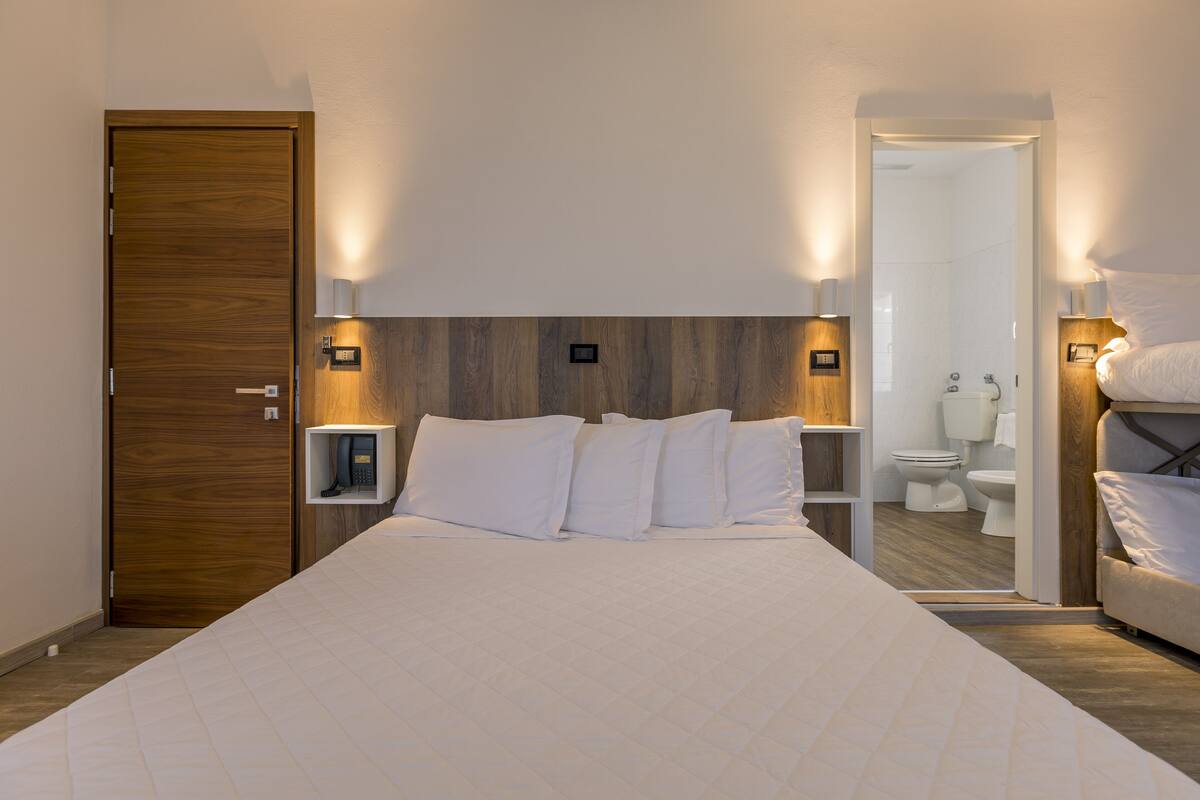 A hotel room with a large bed, wooden headboard, bedside tables with lamps, a built-in shelf, and a mirror.