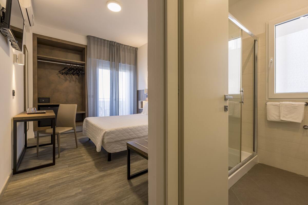 A hotel room with a bed, a desk, a chair, a coat rack, and a shower area.