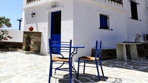 A white building with a blue door and windows, a stone wall, and a tiled patio with blue chairs. - Porto Vlastos (Tinos)