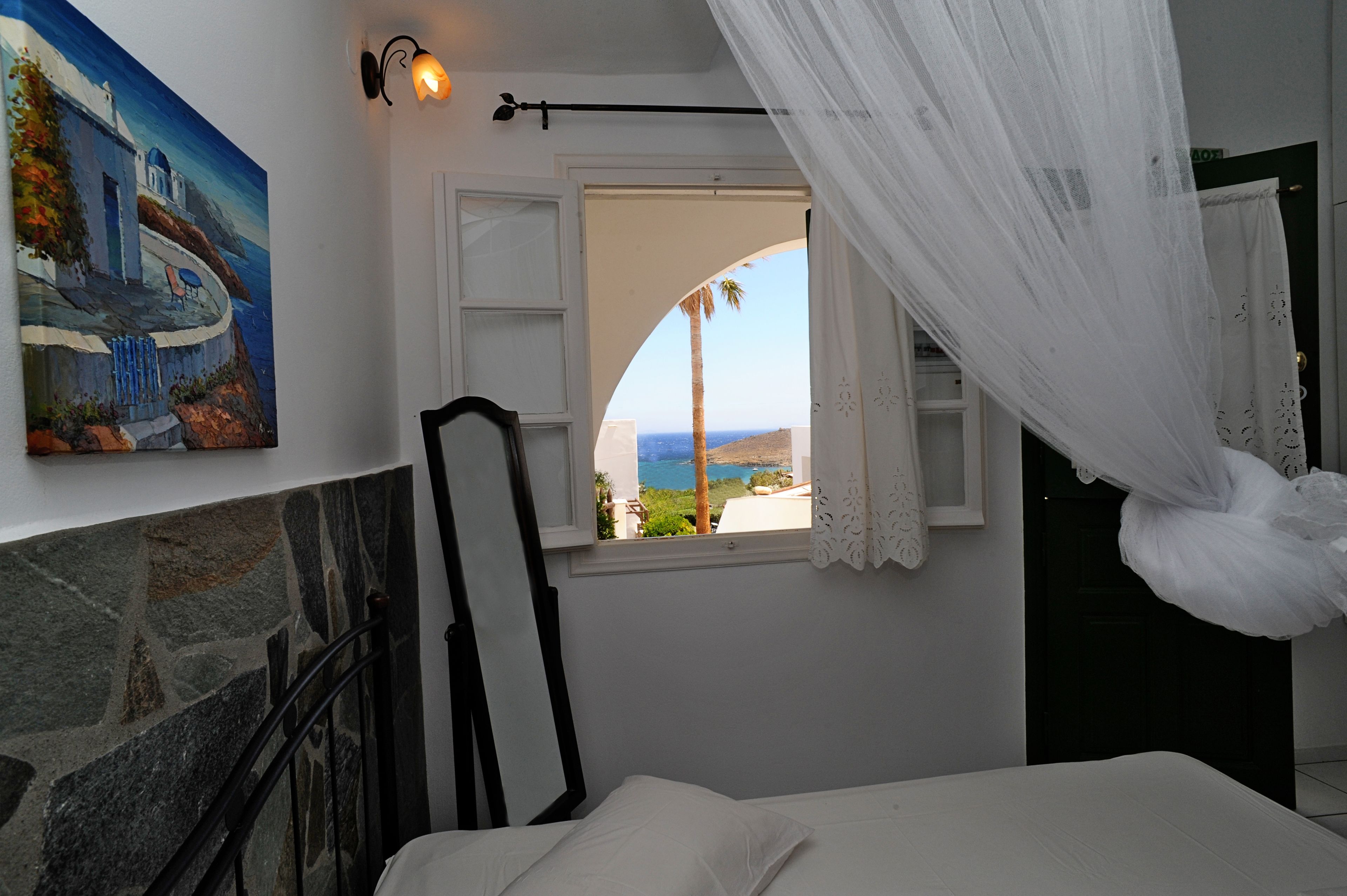 A bedroom with a stone fireplace, a bed with white linens, a painting of a coastal scene, and a window with a view of the sea and palm trees.