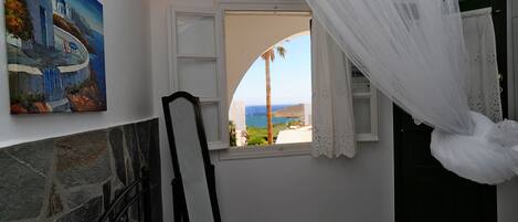 A bedroom with a stone fireplace, a bed with white linens, a painting of a coastal scene, and a window with a view of the sea and palm trees.