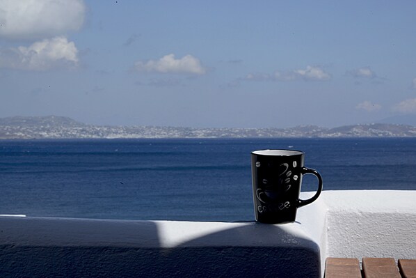 A black mug with a handle on a white ledge overlooking a body of water with distant land and clouds in the sky.