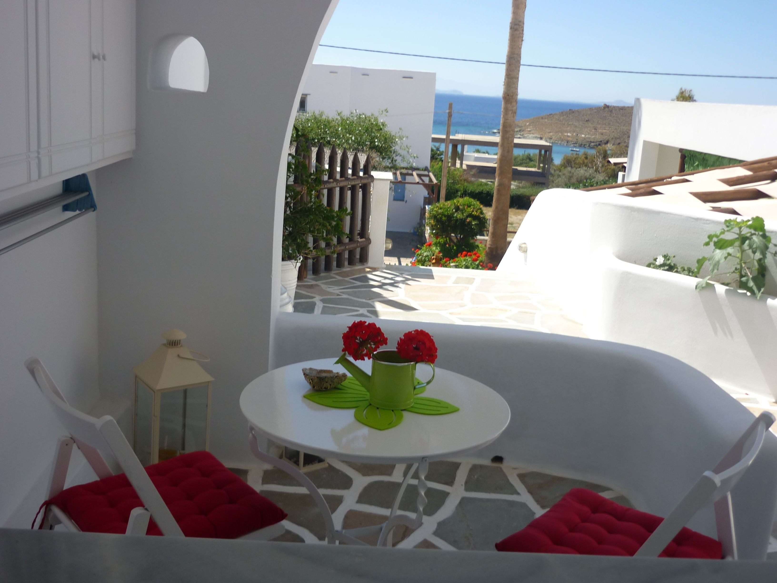 A white room with a table and chairs, a view of the sea, and a white building in the background.