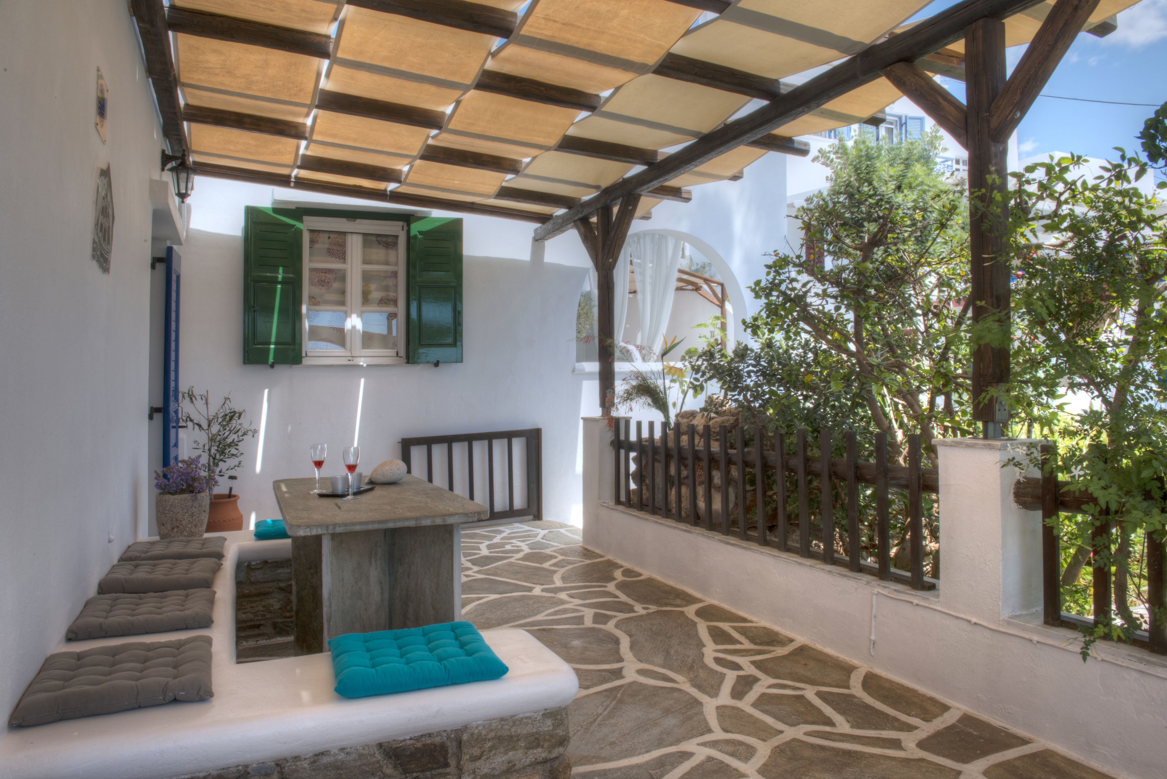 A covered outdoor seating area with a stone floor, a table with cushions, and a view of greenery.