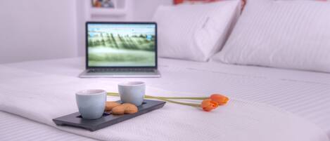 A neatly made bed with a laptop, two cups, cookies, and a flower on a tray.