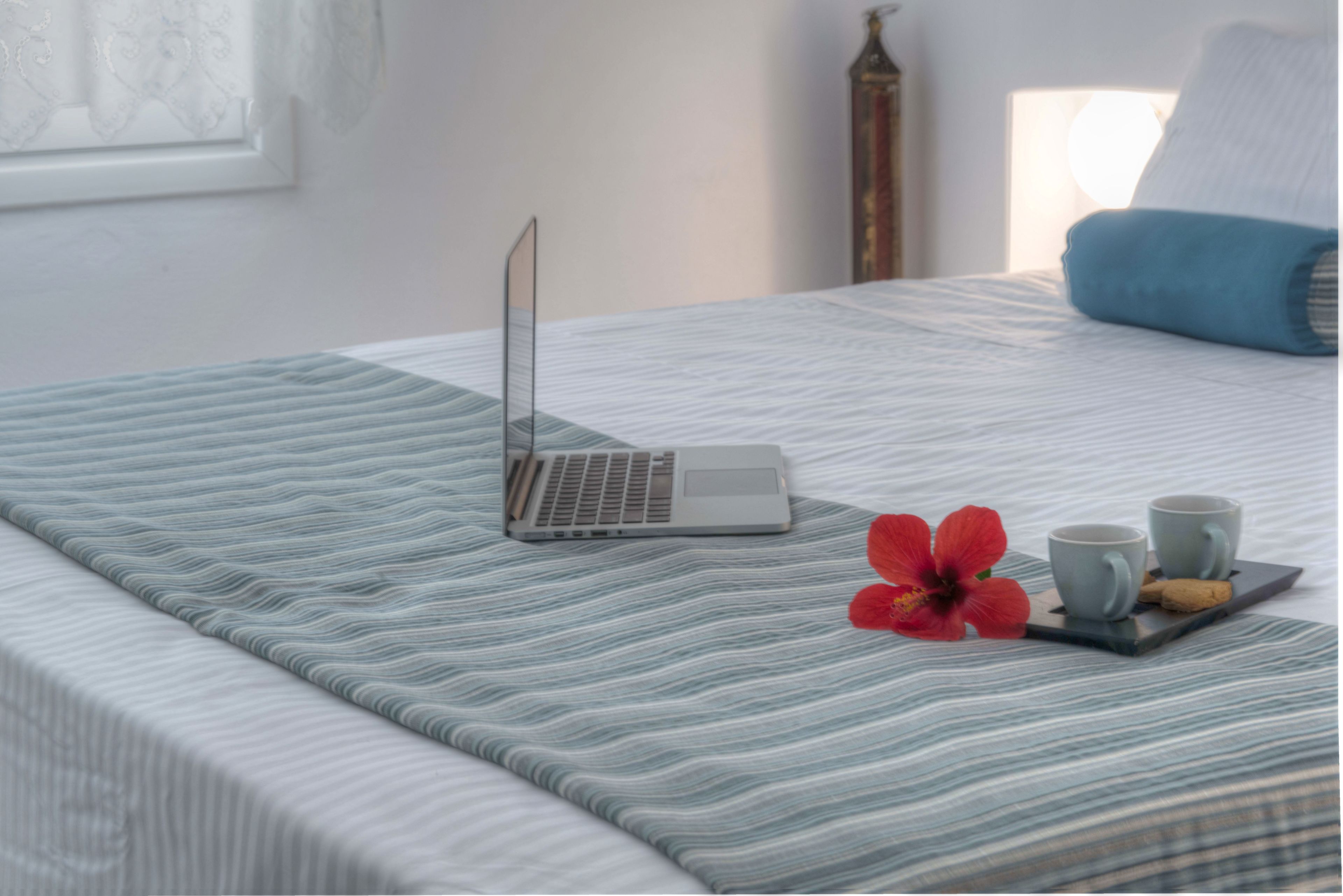 A neatly made bed with a laptop, two cups, and a tray with a cookie and a flower.