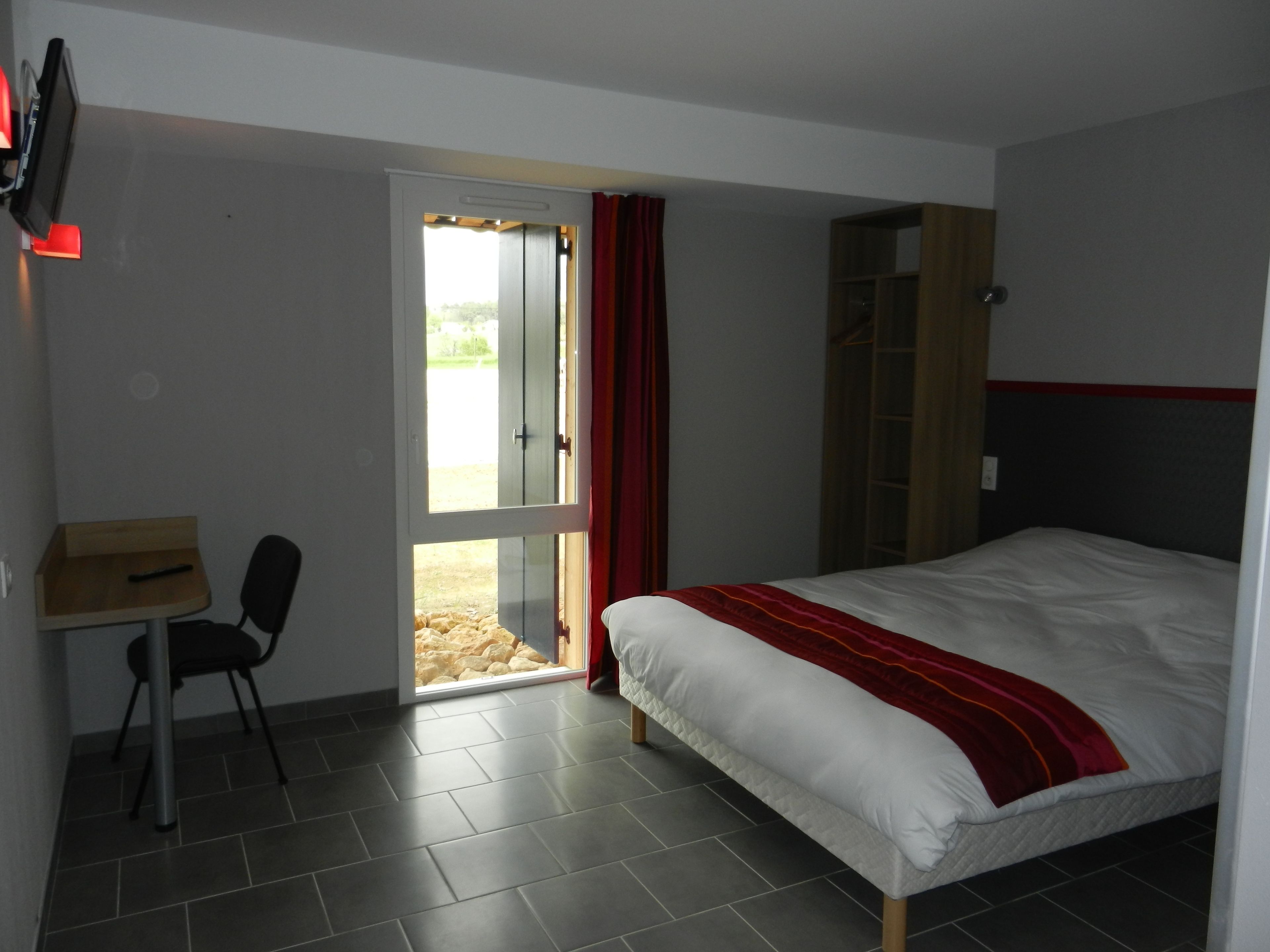 Un dormitorio con cama, escritorio, silla y puerta corredera que da a un área exterior.