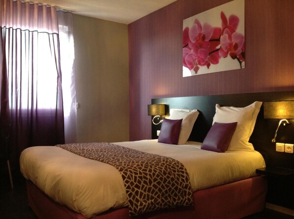 A hotel room with a large bed, a headboard with a light fixture, a wall-mounted painting of pink flowers, and a window with curtains. - The Originals City, Hotel des Lys, Dreux (Dreux)