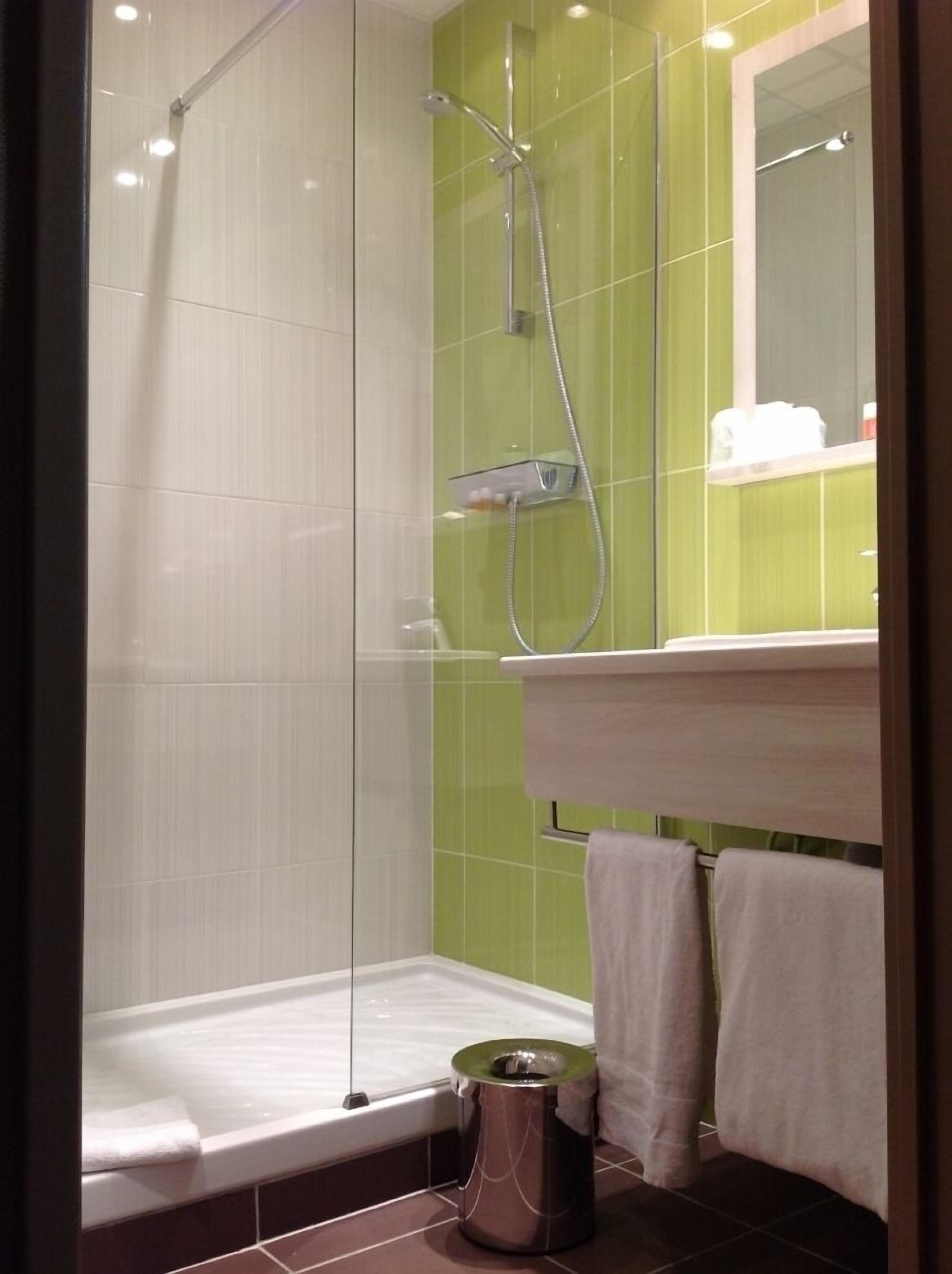 Ducha con puerta de cristal, pared con azulejos verdes, bañera blanca e lavamanos con espejo arriba.