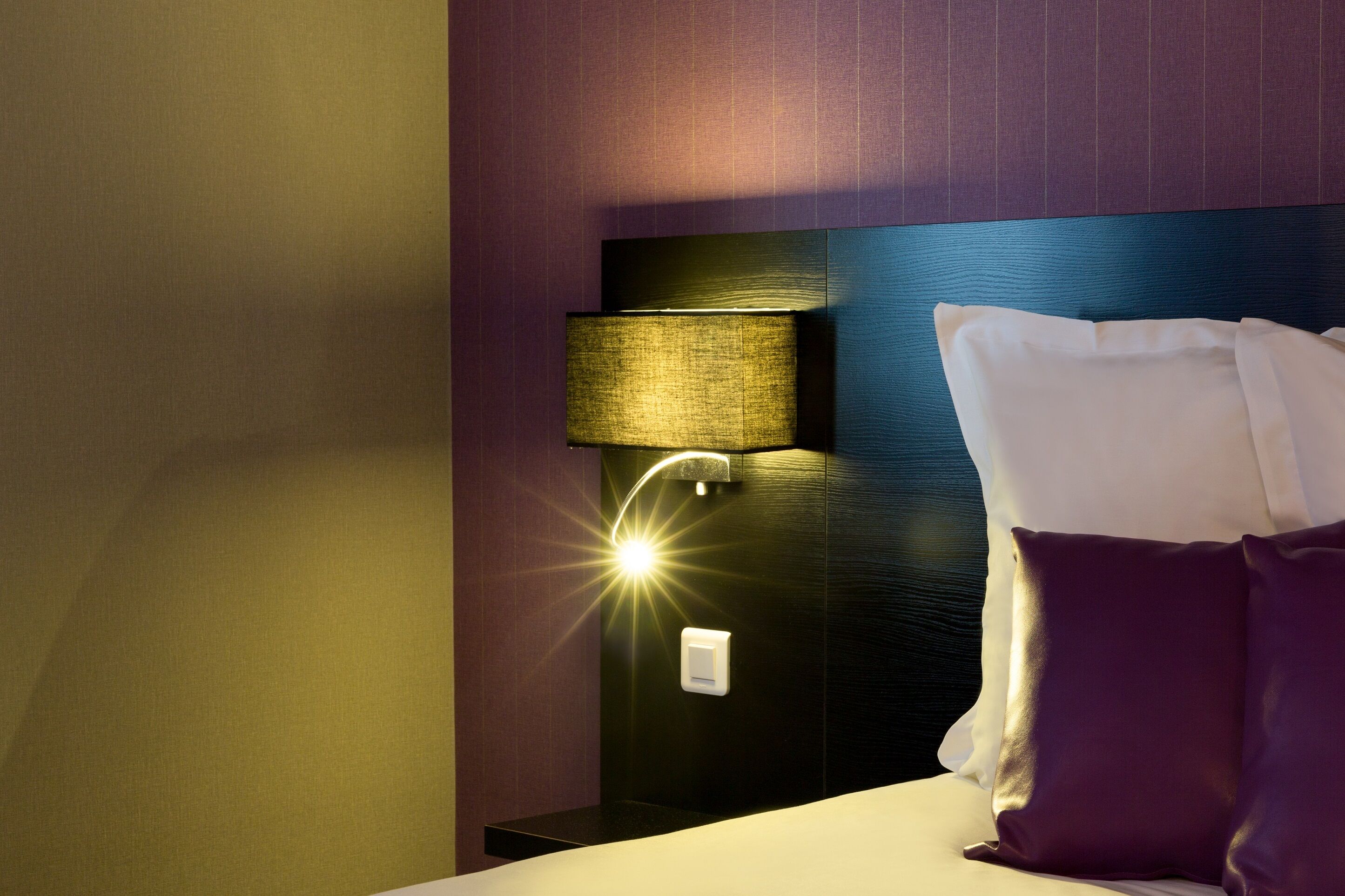 A hotel room with a bed, a nightstand, a lamp, and a wall-mounted light fixture.