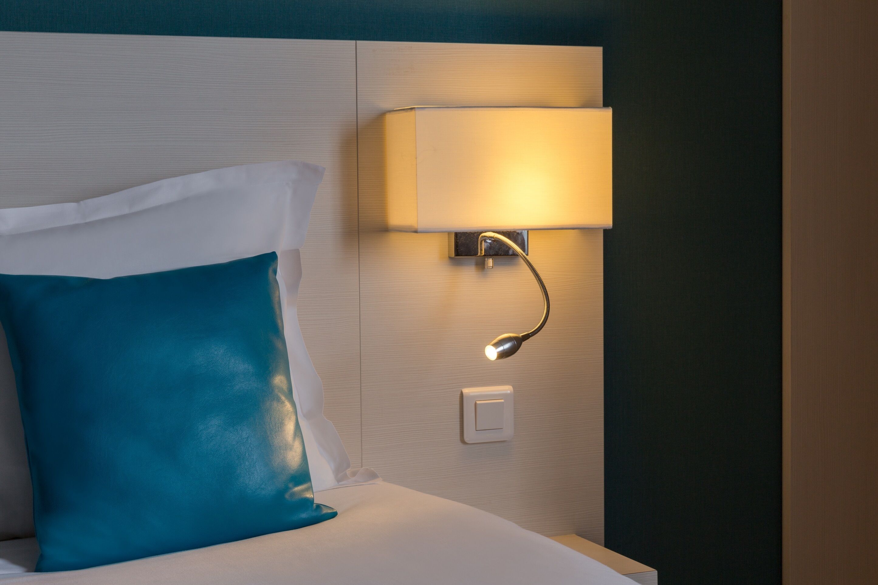 A neatly made bed with a teal pillow and a white pillow, a wall-mounted lamp, and a light switch.