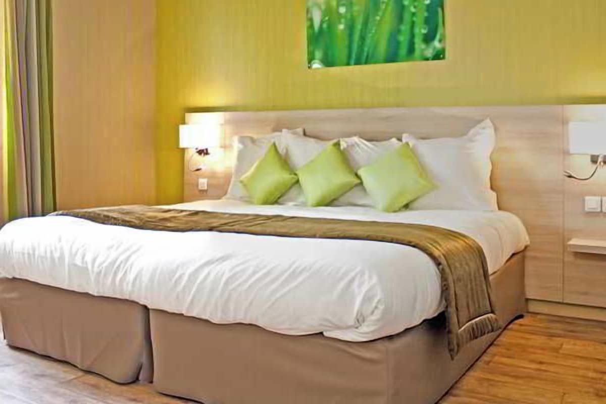 A neatly made bed with a white and beige bedspread, green pillows, and a brown throw blanket.