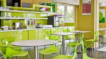 A brightly lit cafeteria with green chairs and tables, a counter with food items, and a window with greenery outside.