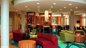 A spacious lobby with a red sofa, yellow armchairs, and a wooden coffee table. There are multiple tables and chairs, a large plant, and a television mounted on the wall. - SpringHill Suites Las Vegas North Speedway (North Las Vegas)