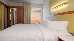 A hotel room with a large bed, a bedside table, a mirror, a window with a view of the sunset, and a wall-mounted light fixture. - SpringHill Suites Las Vegas North Speedway (North Las Vegas)