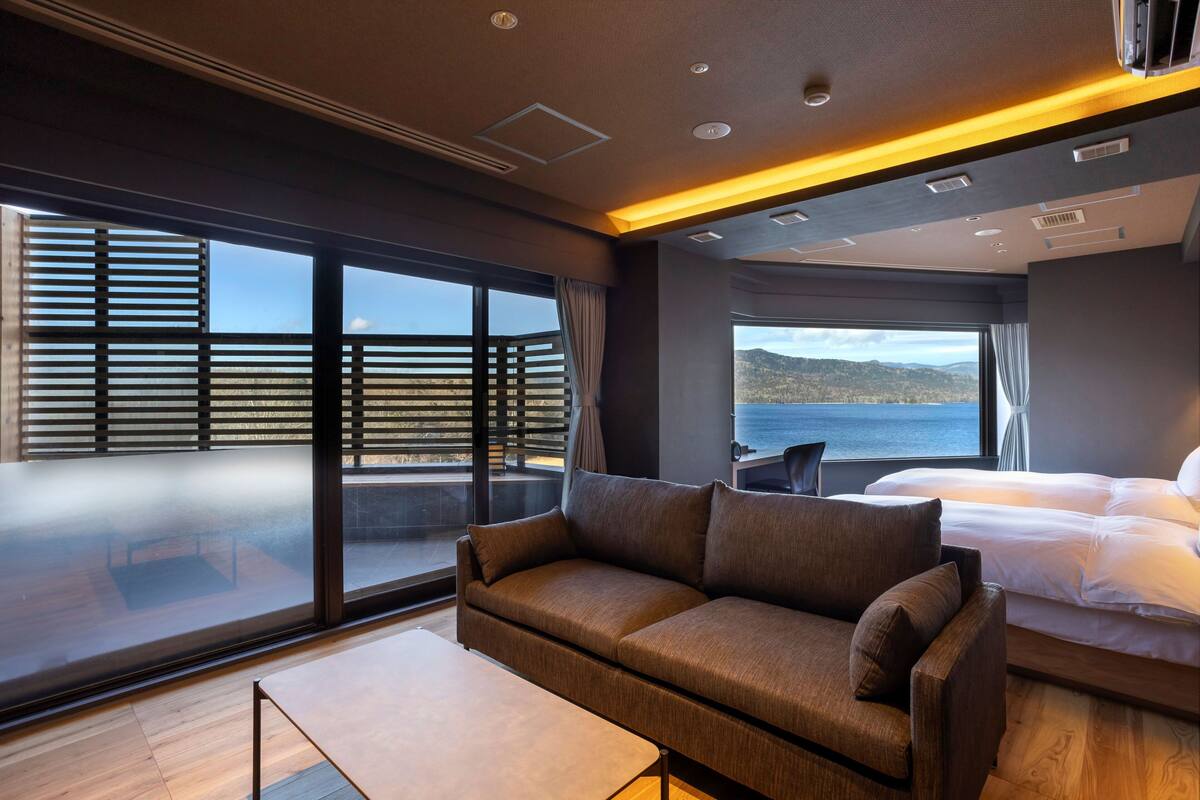 A modern hotel room with a large window, a sofa, a bed, and a small table.