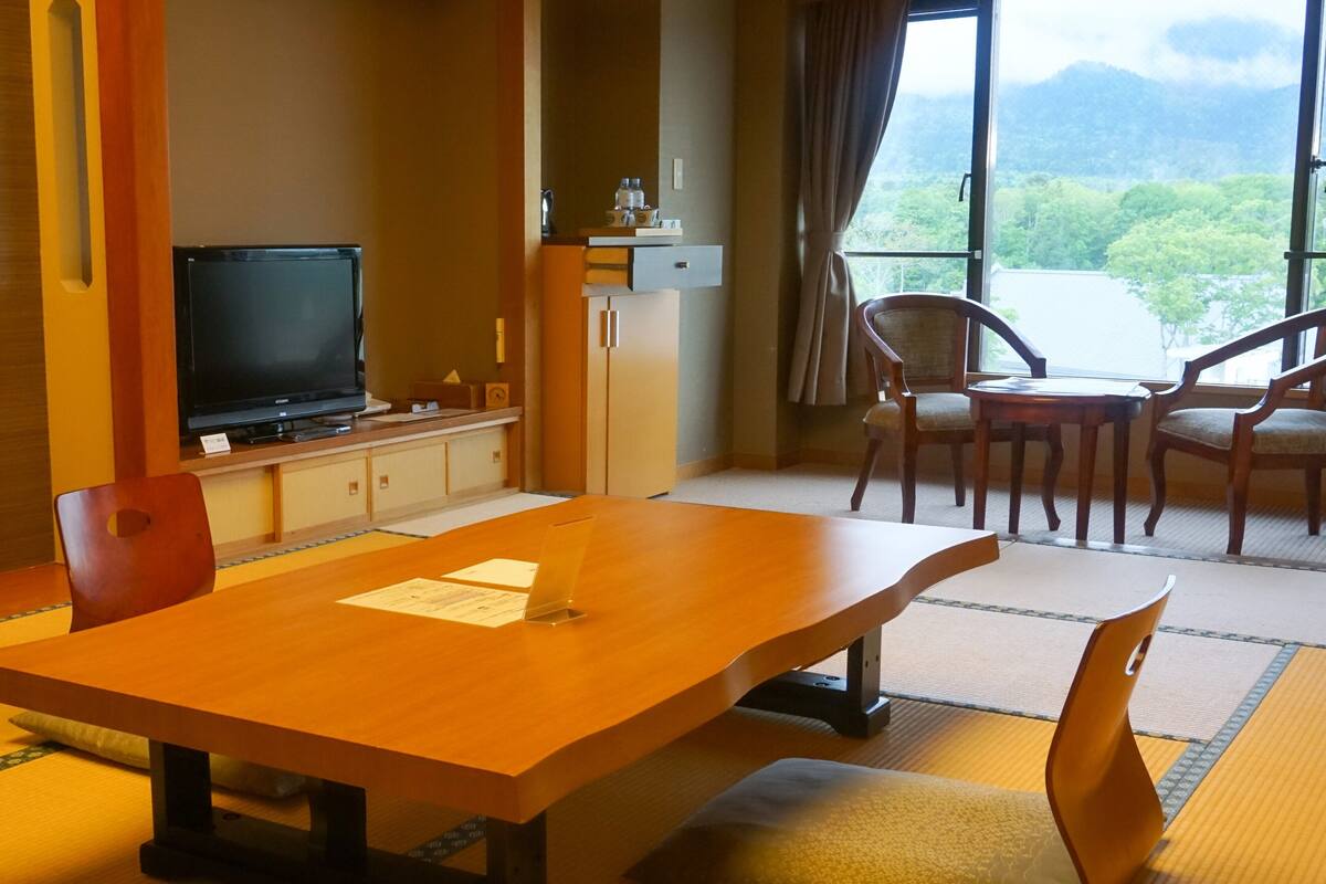A room with tatami flooring, a wooden table, chairs, a television, a small cabinet, and a view of greenery through large windows.