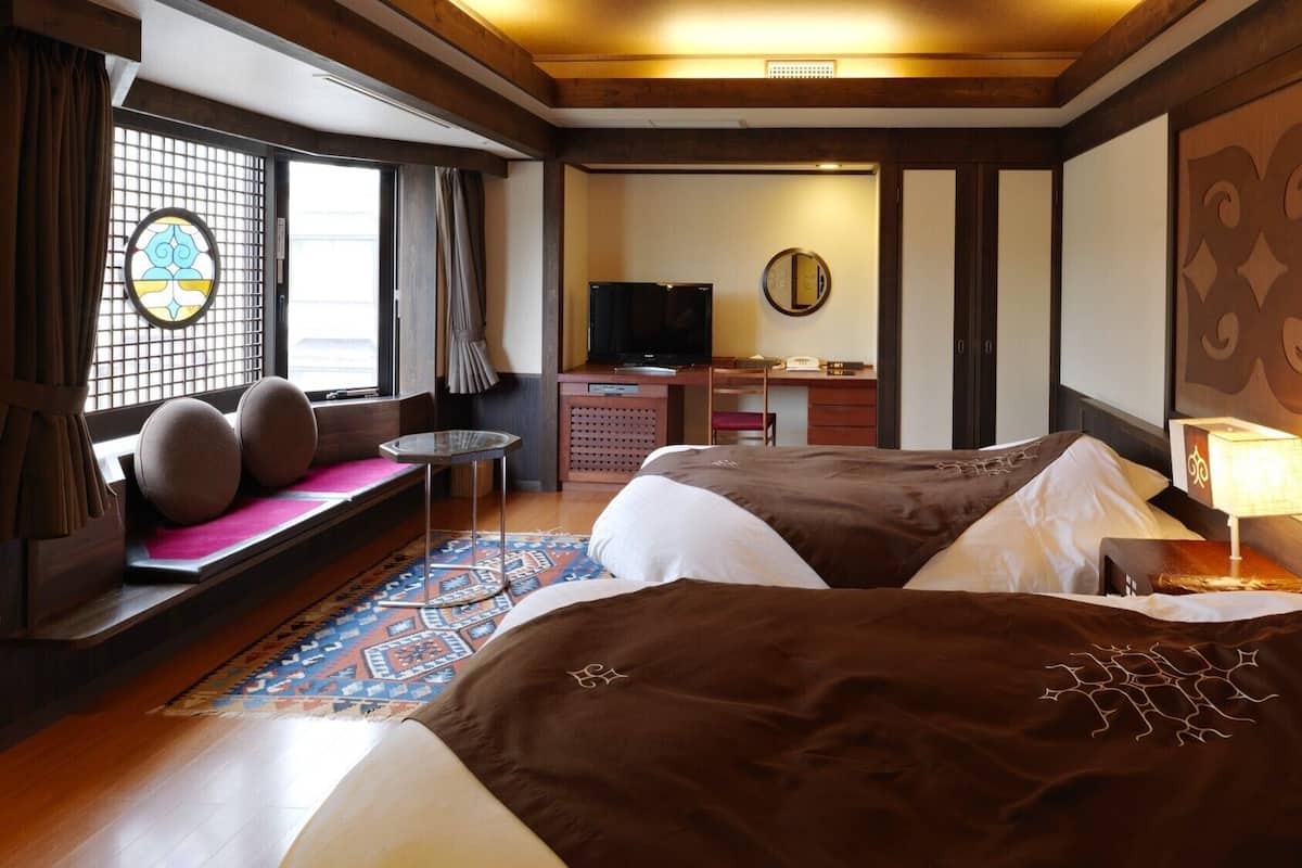 A hotel room with two beds, a desk, a television, a window with a view, and a traditional Japanese-style wall decoration.
