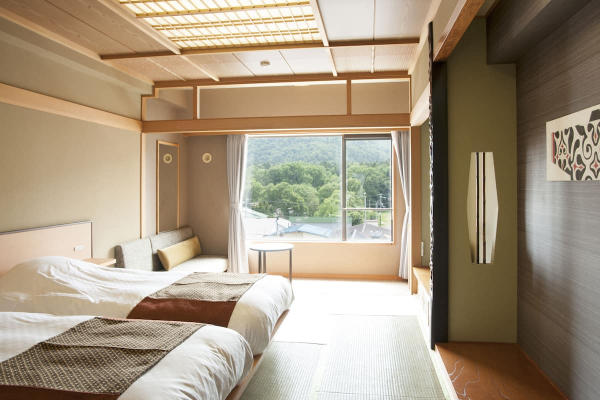 A hotel room with two beds, a large window with a view of greenery, a small table, and a wall-mounted artwork.