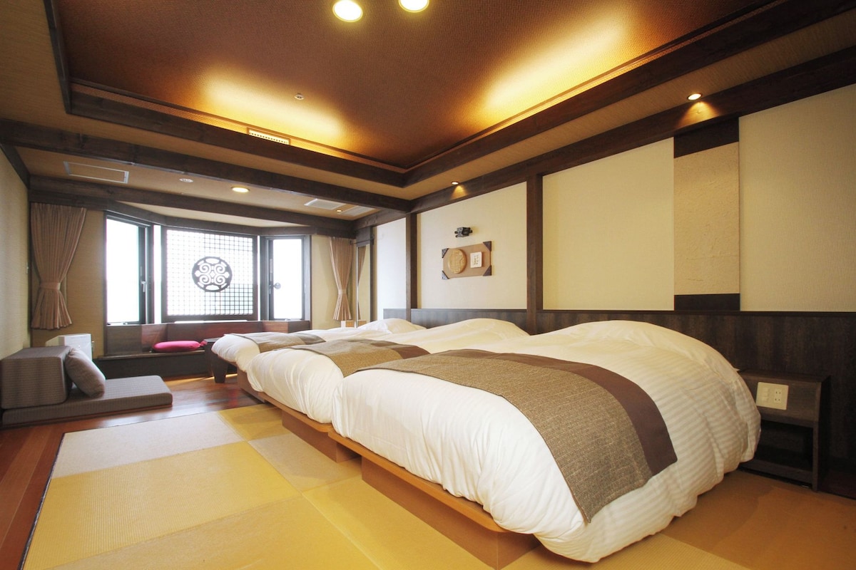 Habitación de hotel de estilo japonés tradicional, con suelo de tatami, una cama grande con muchas almohadas, un futón y una puerta corredera con un ventanal decorativo.