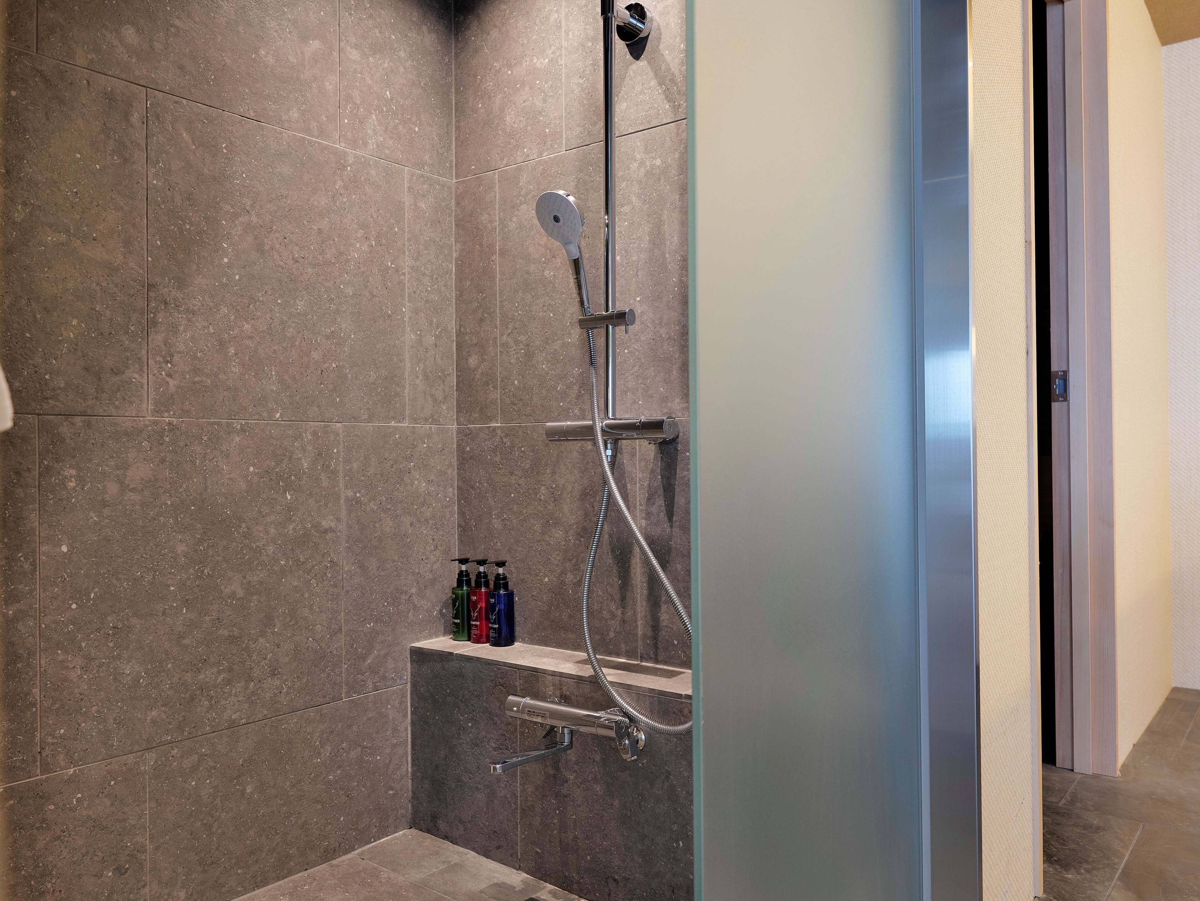 A shower area with a handheld showerhead, a wall-mounted faucet, and a handheld shower attachment. There are three bottles of shampoo on a shelf below the showerhead.