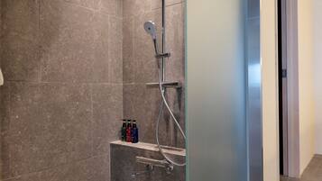A shower area with a handheld showerhead, a wall-mounted faucet, and a handheld shower attachment. There are three bottles of shampoo on a shelf below the showerhead.