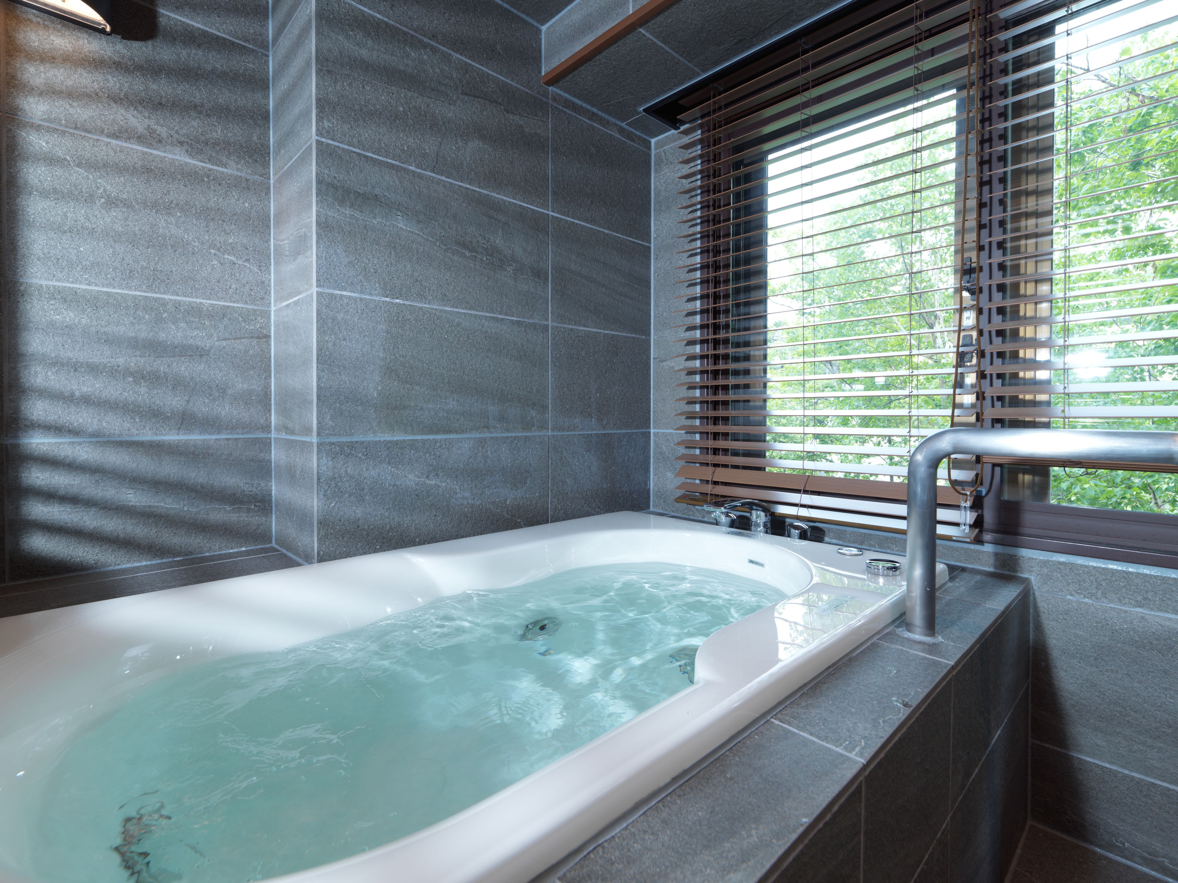 A modern bathroom with a freestanding bathtub, tiled walls, and a window with blinds.