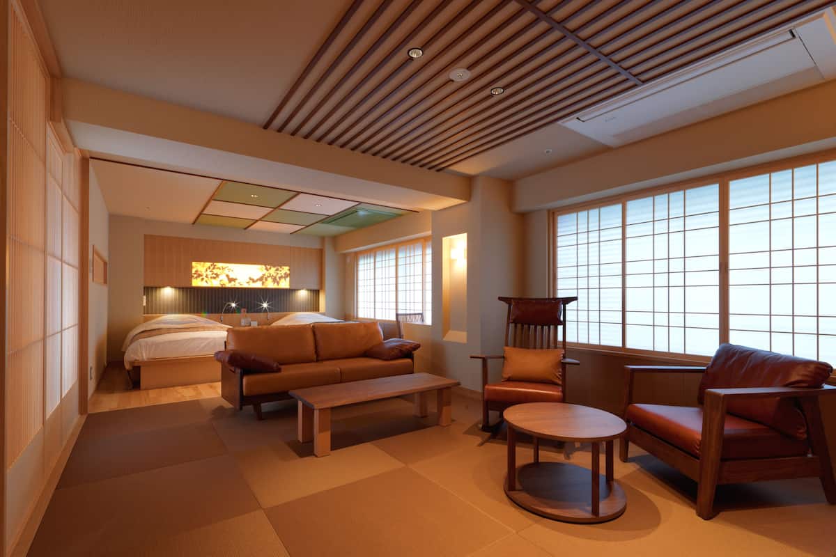 A spacious room with a bed, two sofas, a coffee table, and a chair. There is a large window providing natural light.