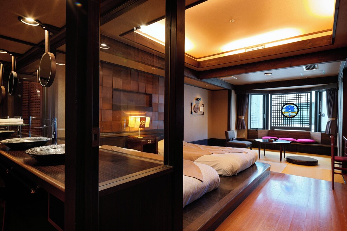 Habitación de hotel con piso de madera, cama con almohadas, una mesita, una silla y ventana con vistas.