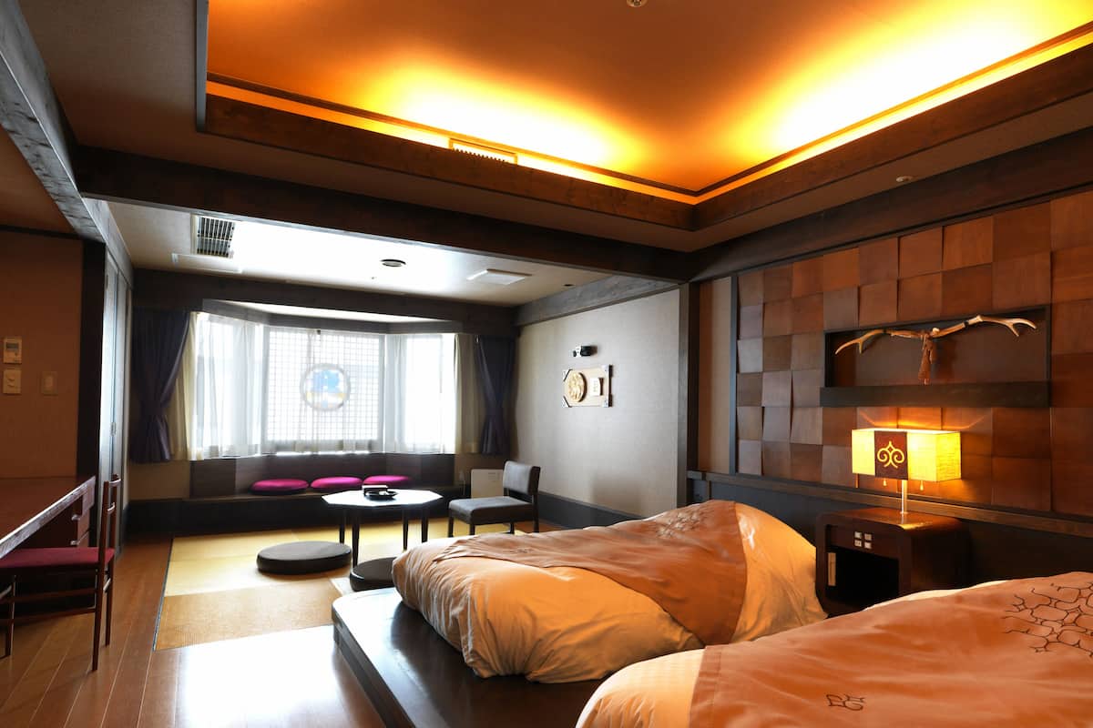 A hotel room with two beds, a small table, a chair, a sofa, and a window with curtains.