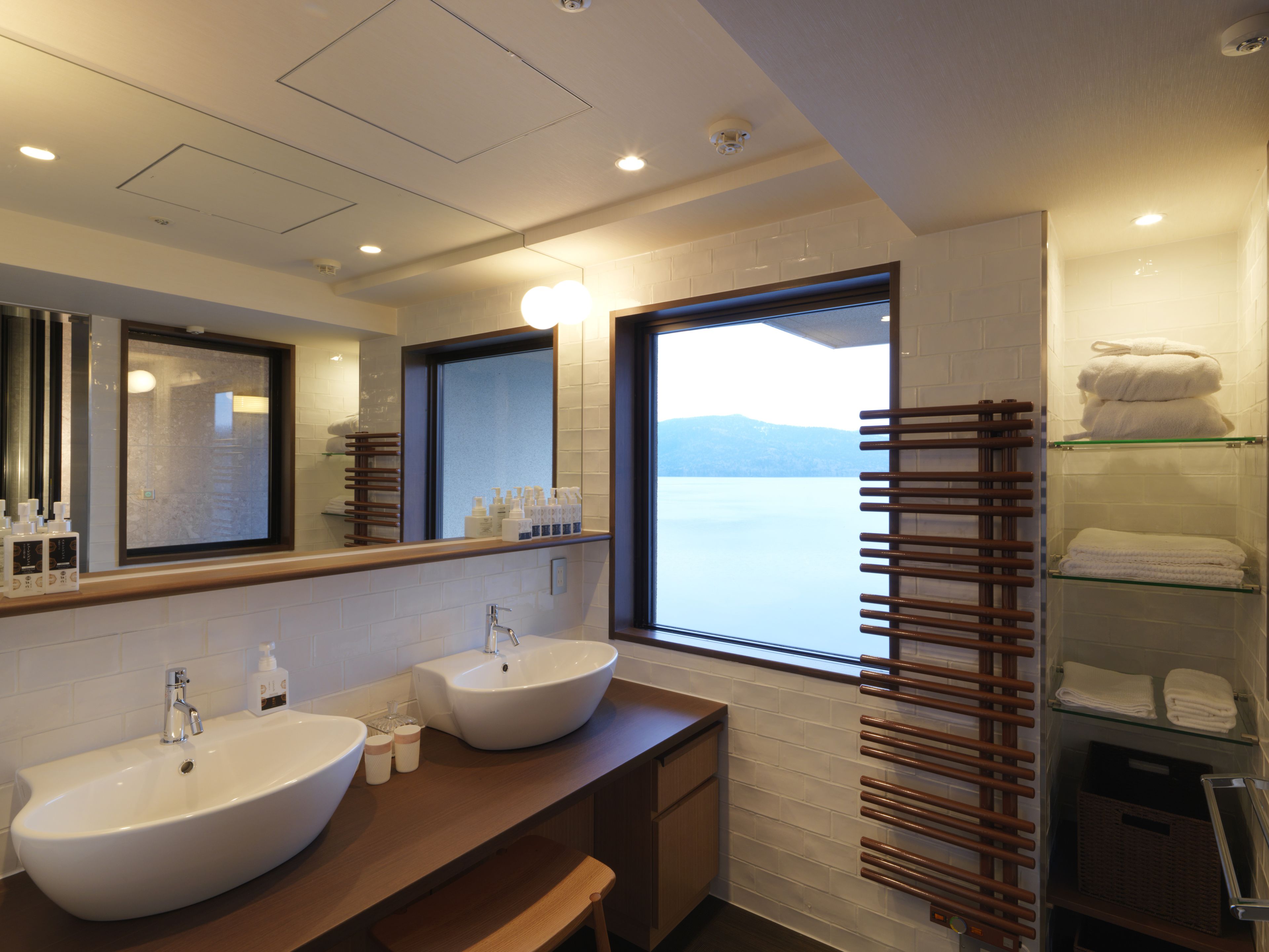 A modern bathroom with two sinks, a large mirror, and a window with a view of a lake.