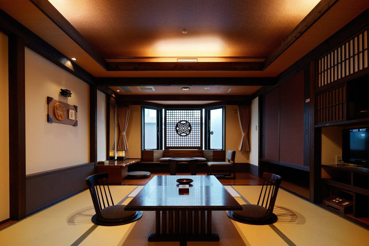A traditional Japanese room with tatami flooring, a low table with chairs, a sliding door with a decorative screen, and a television on a wooden stand.