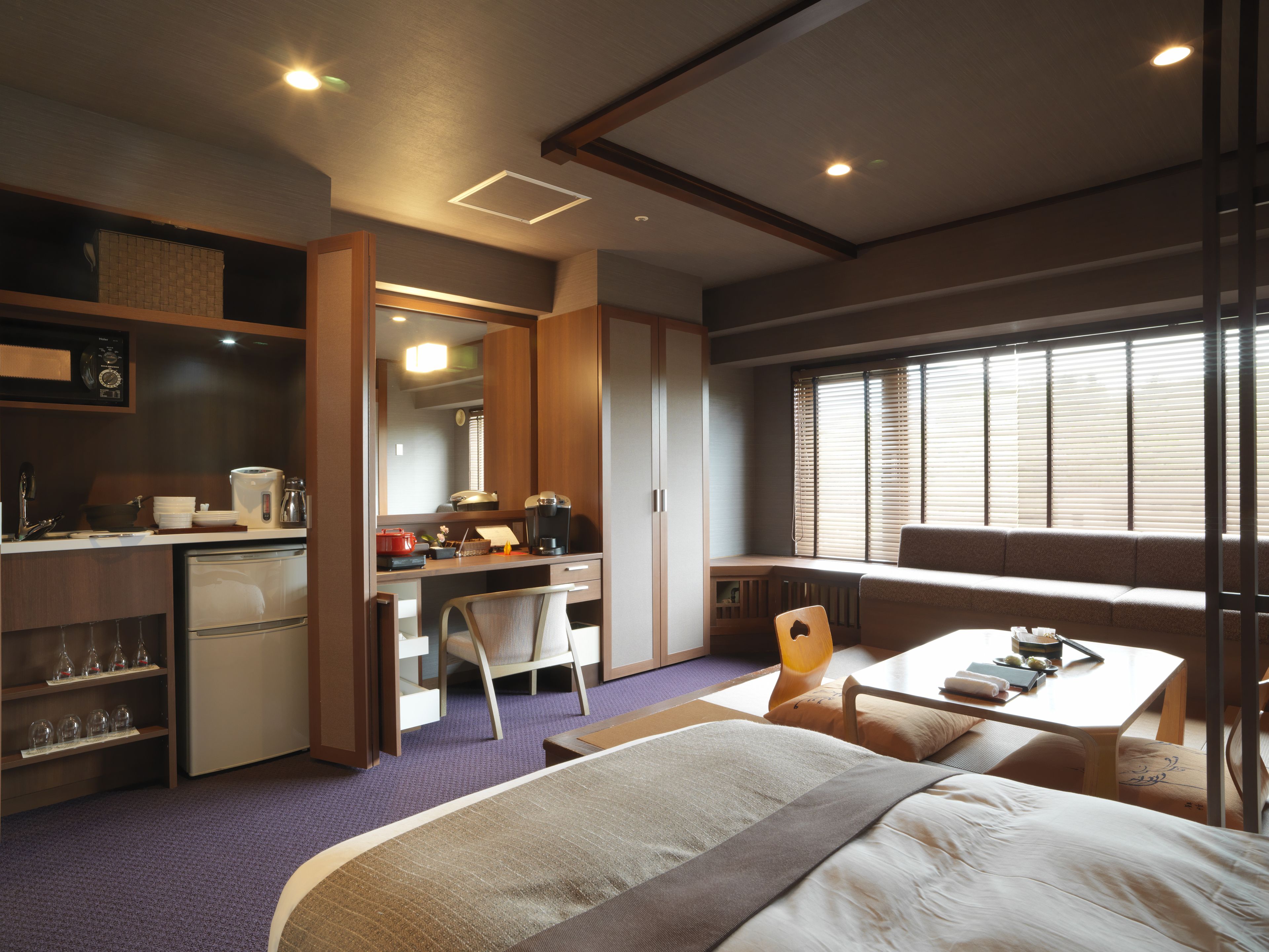 A compact hotel room with a kitchenette, a bed, a dining area, and a large window with blinds.