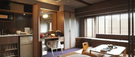 Habitación de hotel compacta con kitchenette, cama, zona de comedor y un amplio ventanal con persianas.