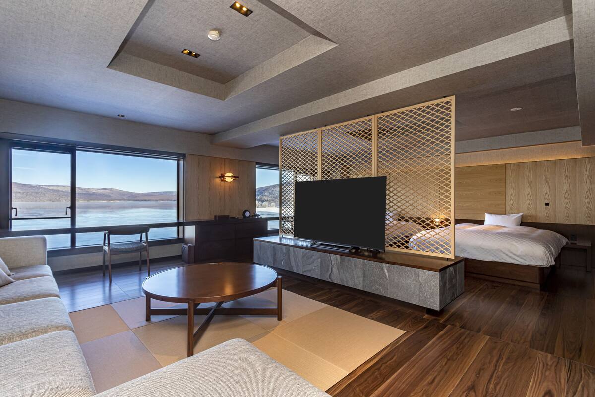 A modern hotel room with a large window, a flat-screen TV, a sofa, a coffee table, and a bed.
