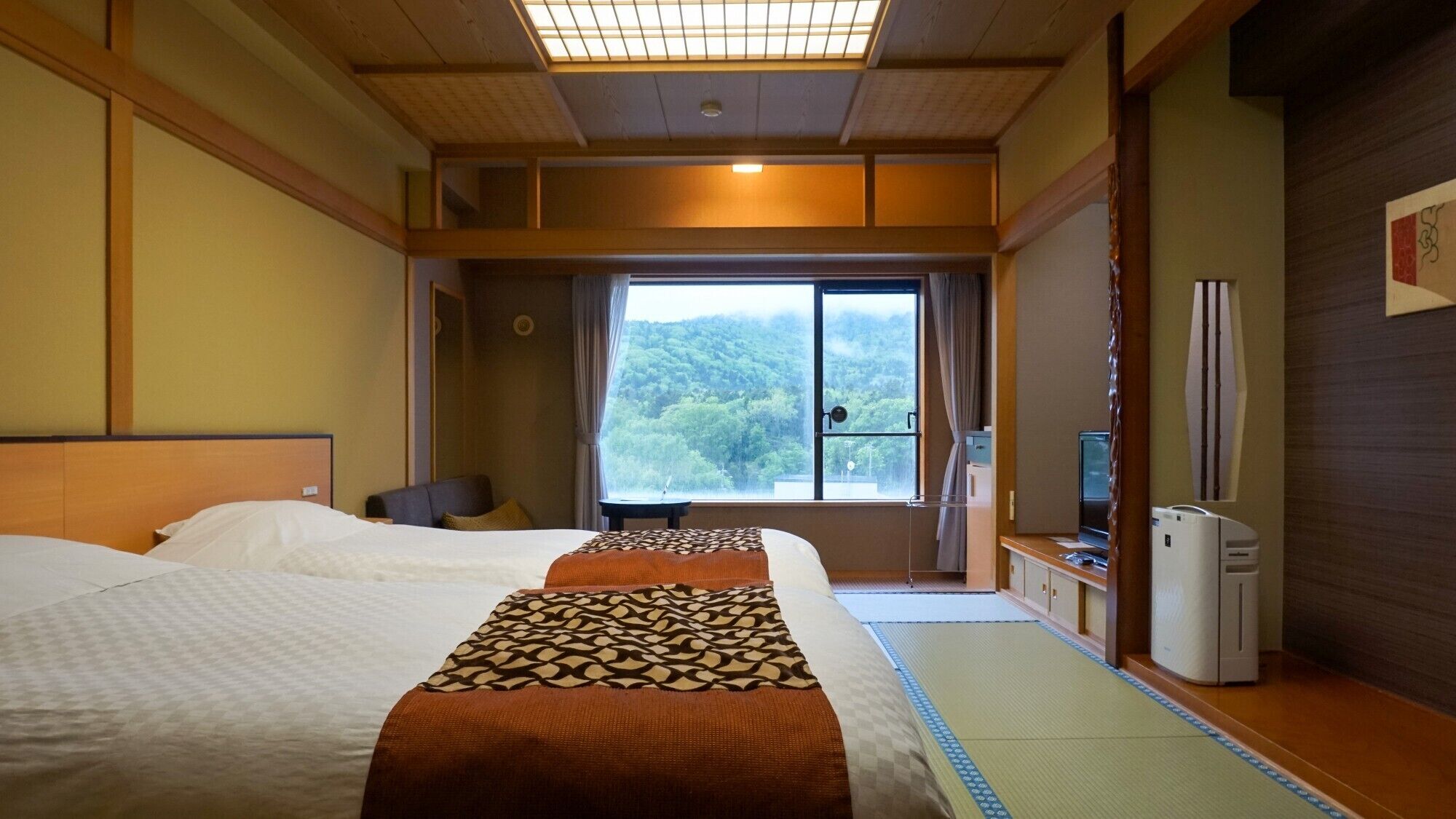 A hotel room with two beds, a desk, a chair, a TV, and a window with a view of a forest.
