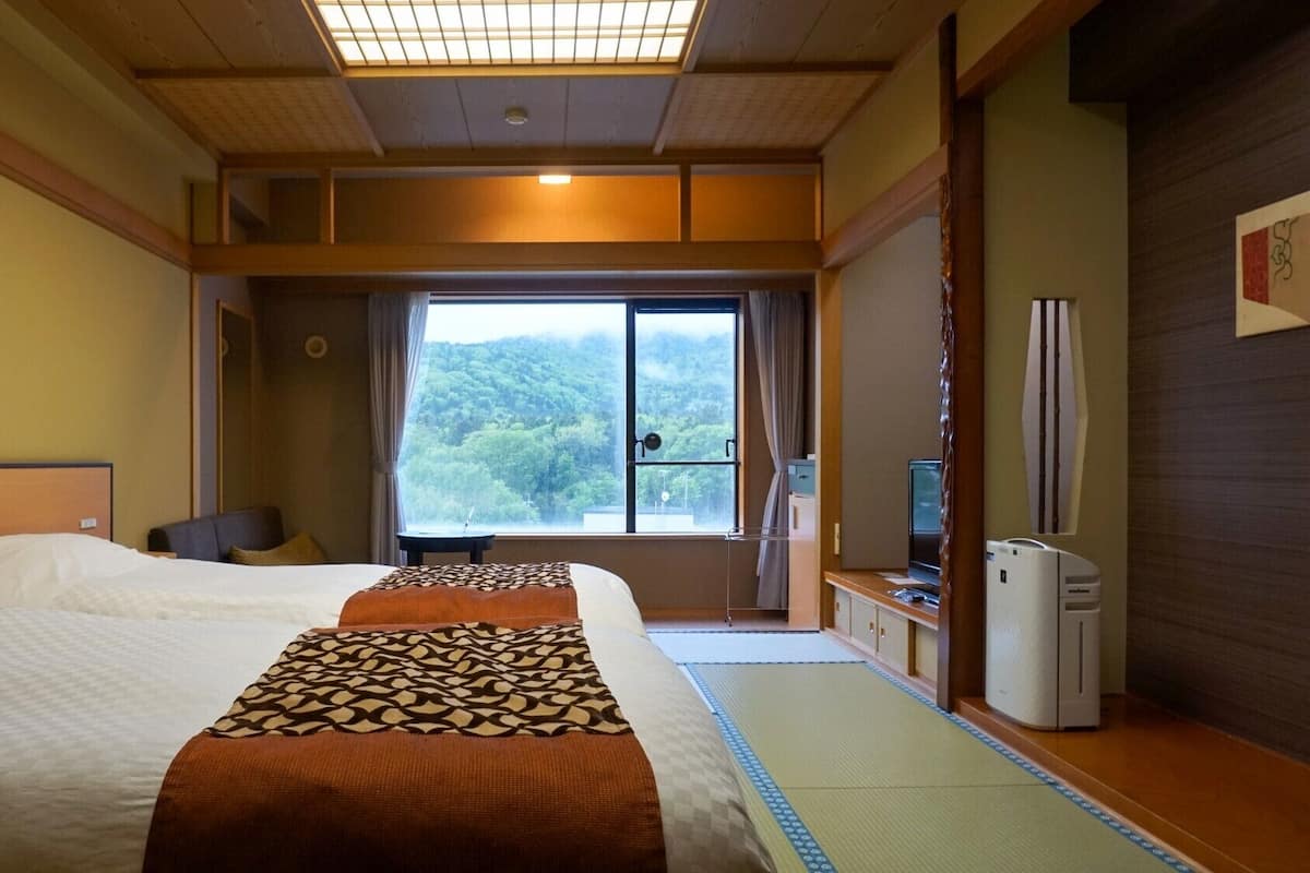 A hotel room with two beds, a desk, a chair, a TV, and a window with a view of a forest.
