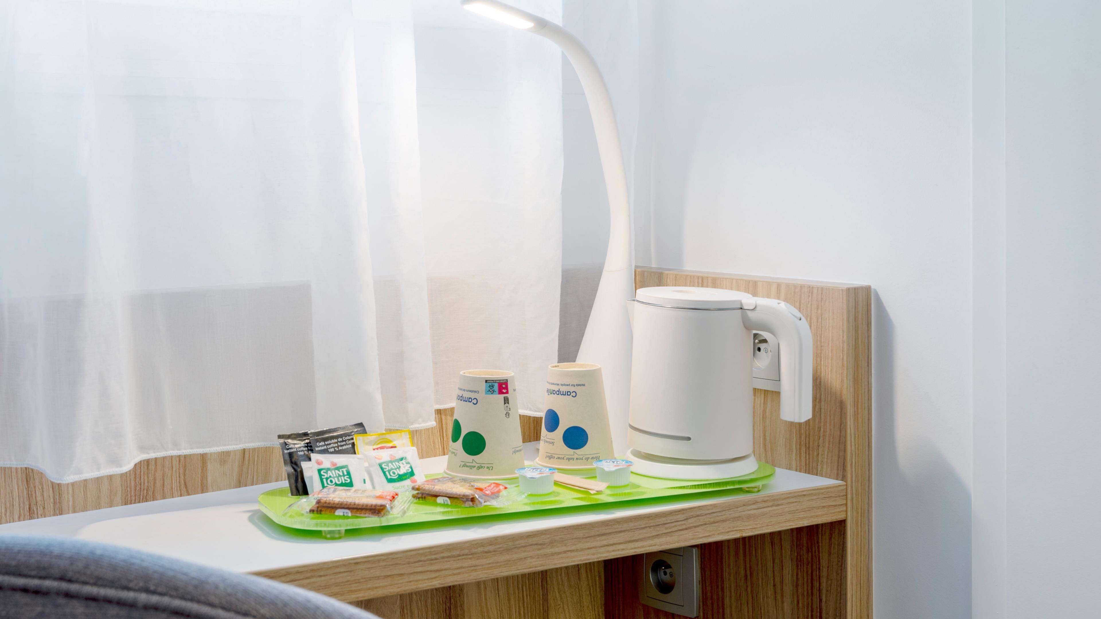 A hotel room with a wooden nightstand, a white kettle, a green tray with snacks, and a white lamp.