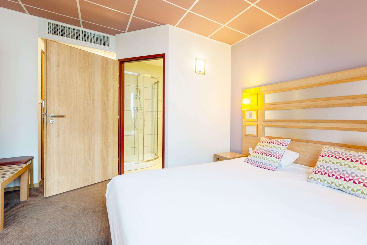 Hotellihuone, jossa on sänky, yöpöytä ja suihkutila.