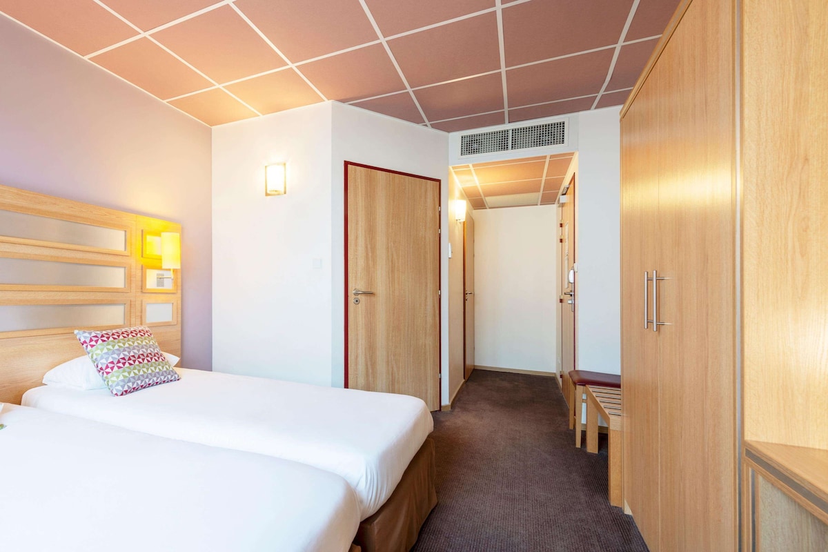 Hotellihuone, jossa on kaksi sänkyä, puinen sängynpääty, pieni työpöytä ja vaatekaappi.