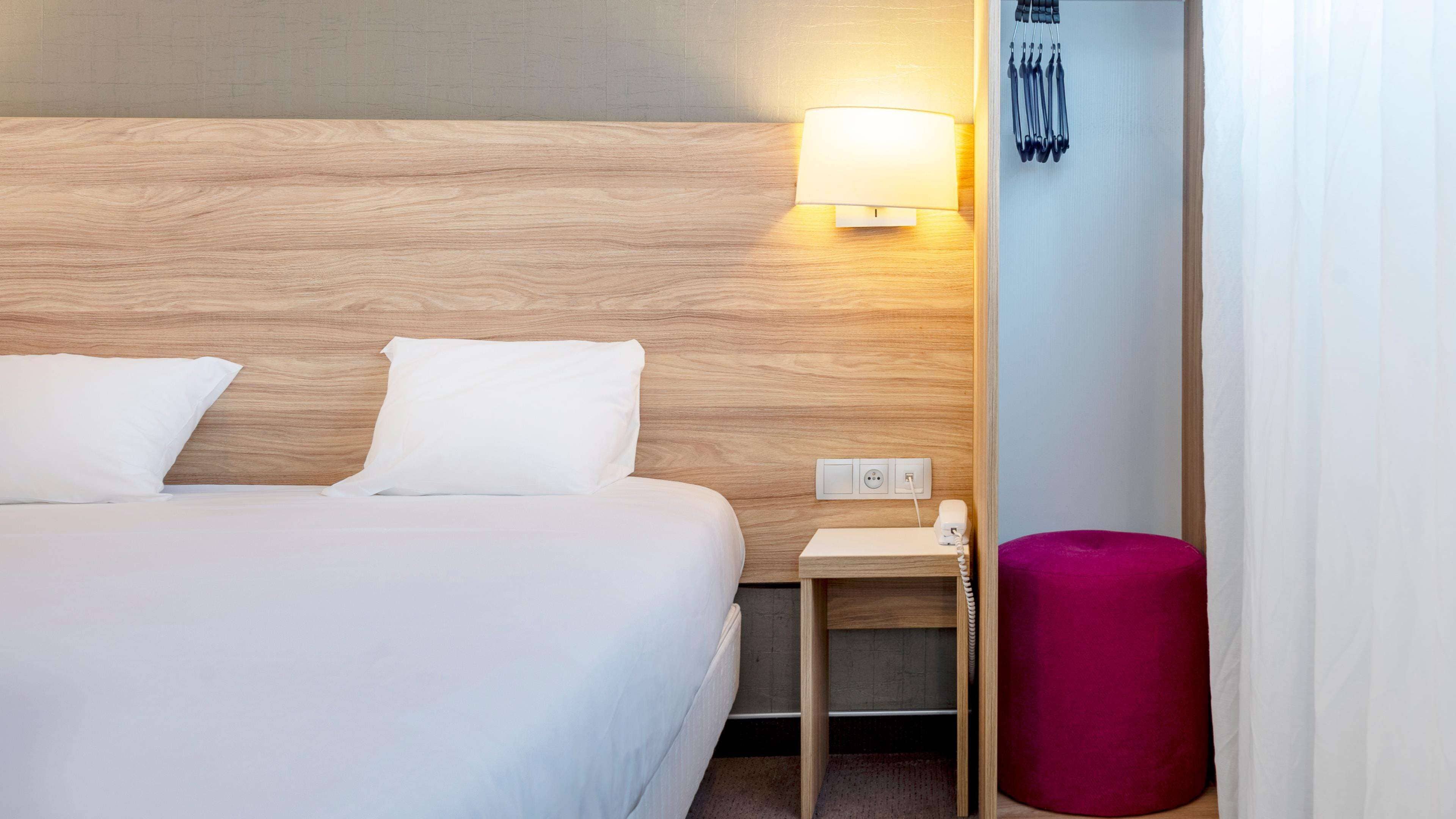 A hotel room with a wooden headboard, a bedside table with a lamp, a phone, and a pink ottoman.