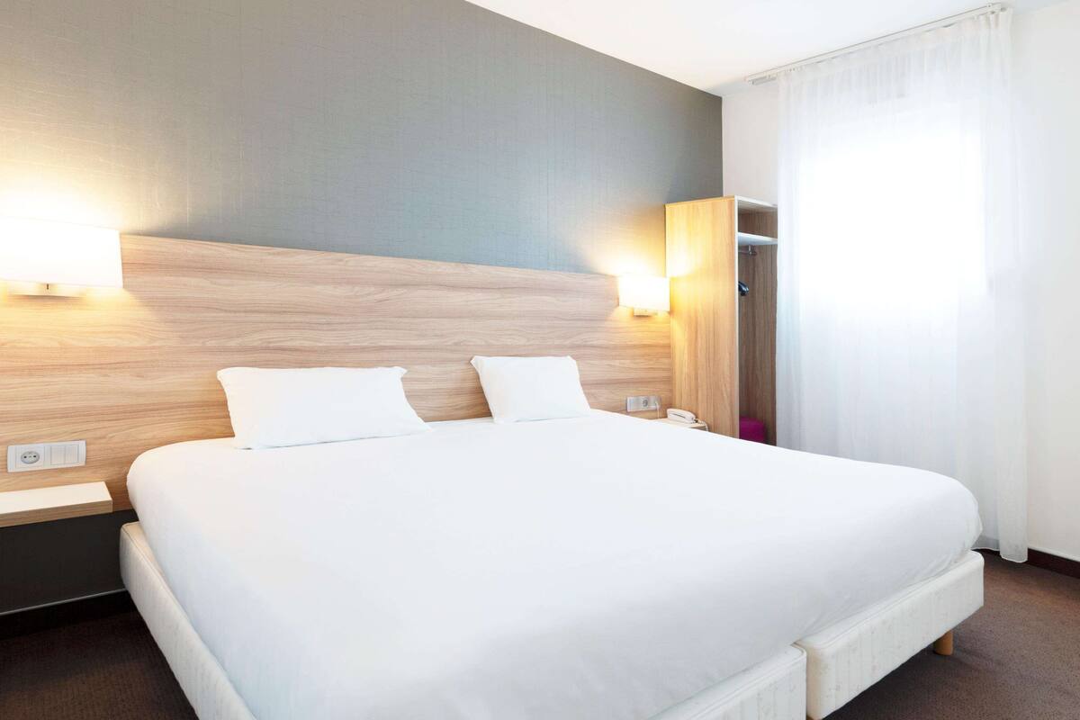 A hotel room with a large bed, a wooden headboard, two bedside lamps, a nightstand, and a red ottoman.